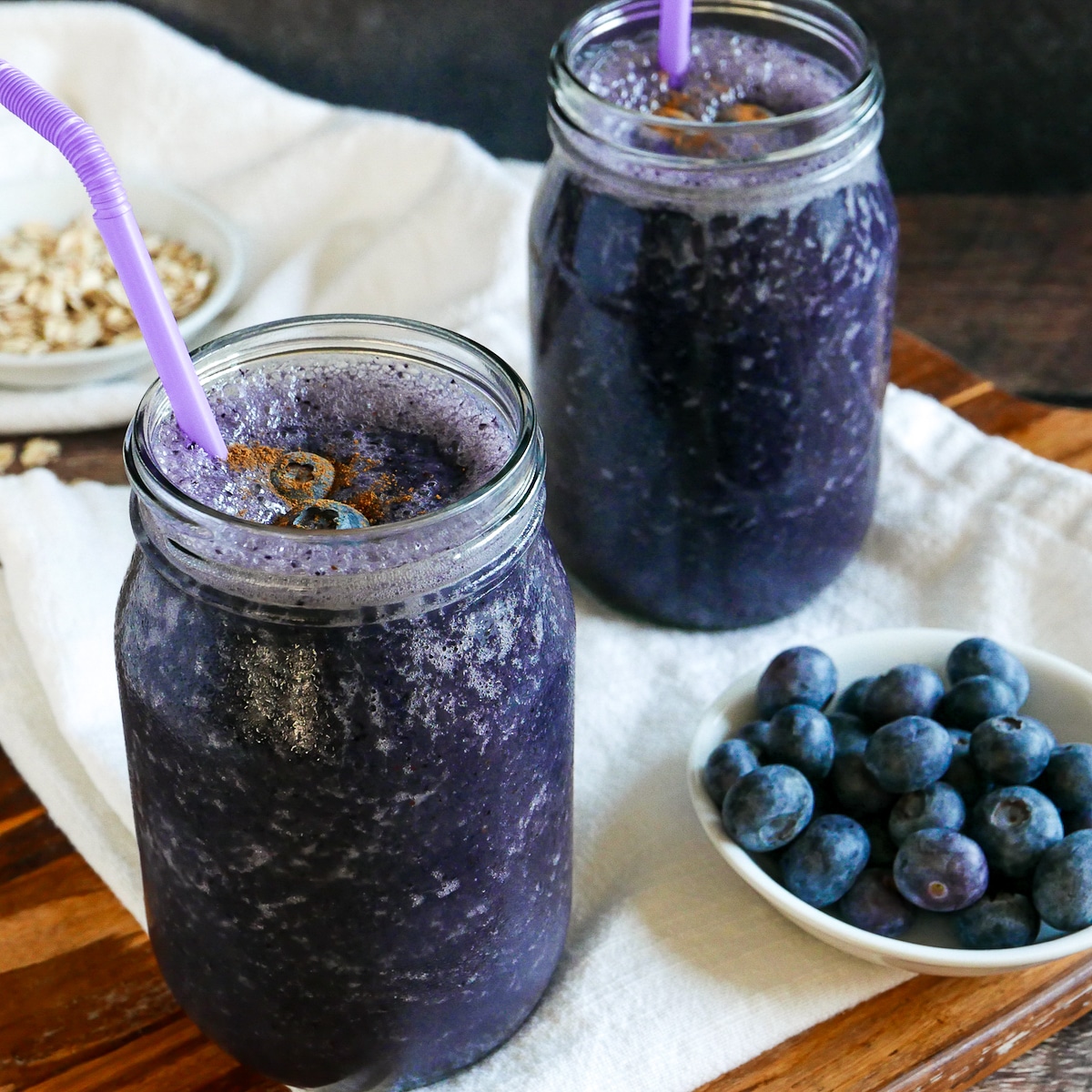 Two oat milk smoothies with blueberries and oats in cups in the background.