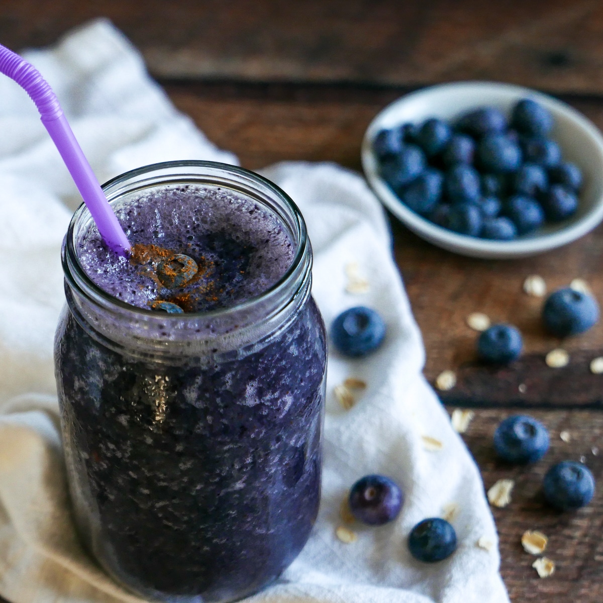 oat-milk-smoothie-with-straw.jpg Oat milk protein shake in a glass jar and a cup of blueberries in the background.