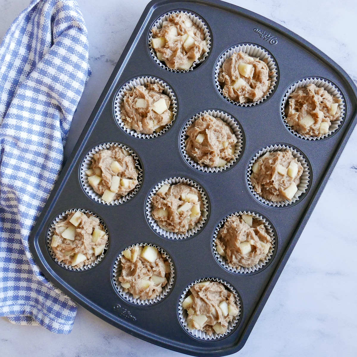 Muffin tin filled with apple muffin batter and a napkin on the side.
