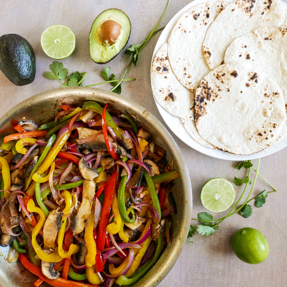 cooked-veggies-and-flour-tortillas.jpg cooked veggies in skillet and flour tortillas on a plate with lime, avocado, and cilantro.