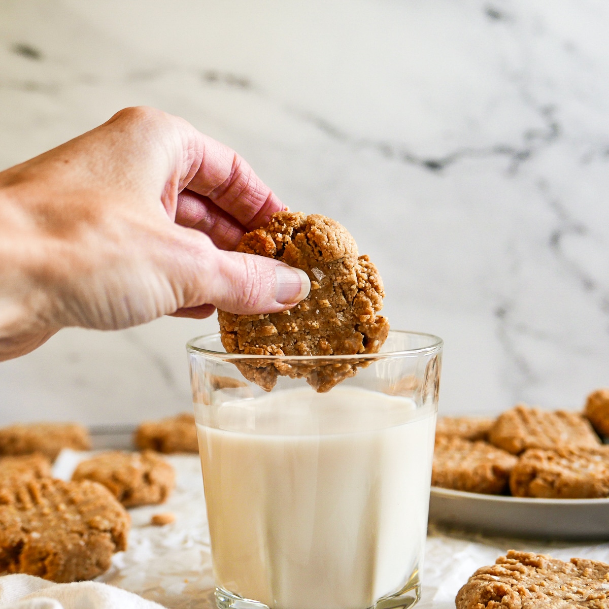 white hand dunking cookie into glass of milk with a plate of cookies in background.