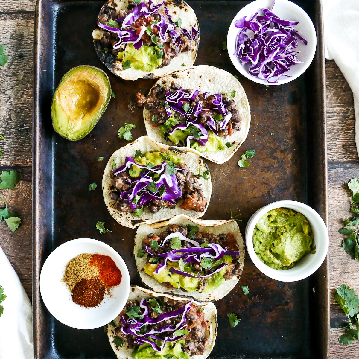 five refried bean tacos arranged on a baking sheet with red cabbage, avocado, and cilantro. 