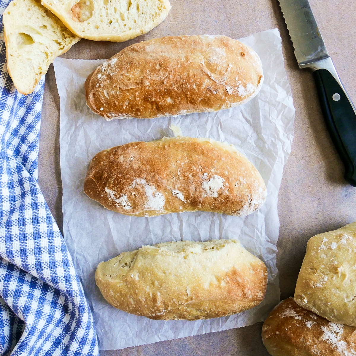 three ciabatta buns arranged on parchment paper with a bread knife next to them.