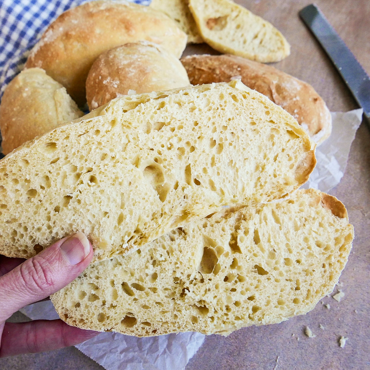 Hand holding up cut open ciabatta roll with more rolls in the background. 