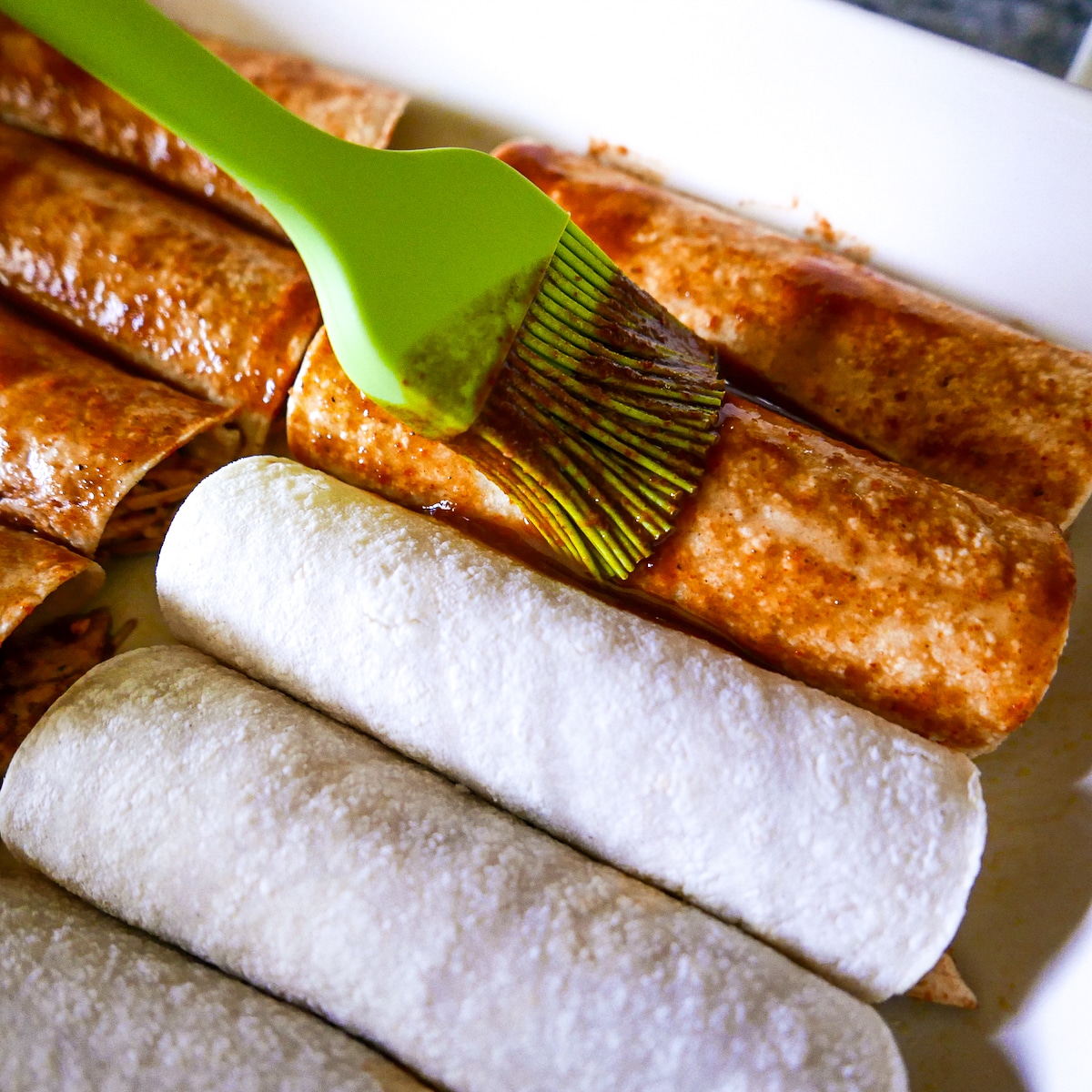 enchilada sauce being brushed on top of prepped enchiladas.