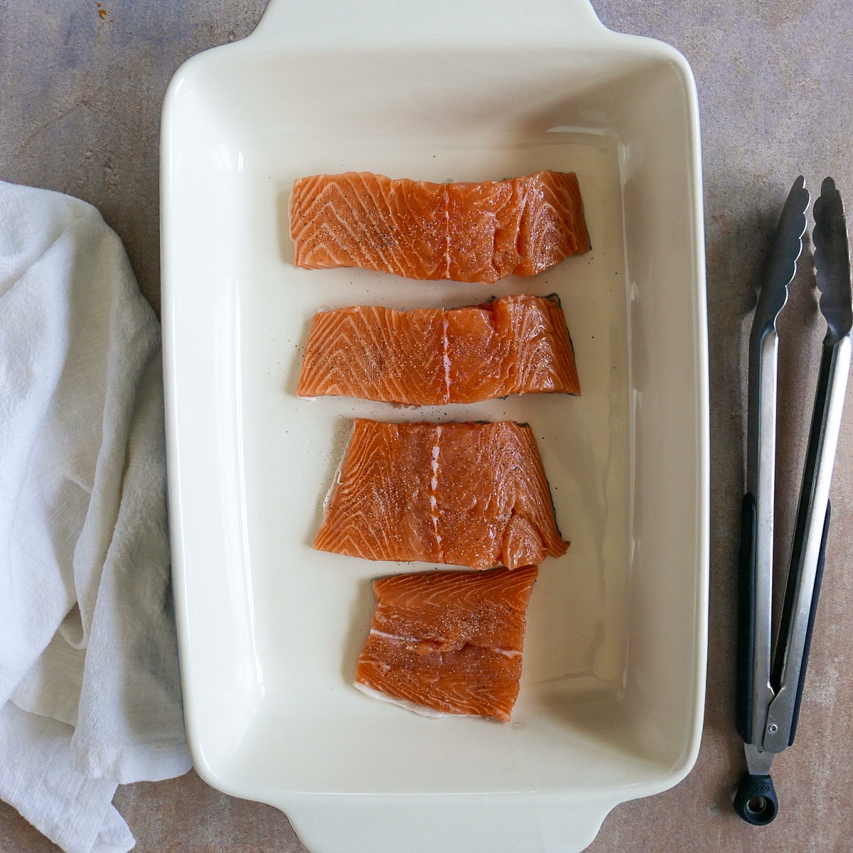 four salmon fillets in a large baking dish with tongs resting nearby.