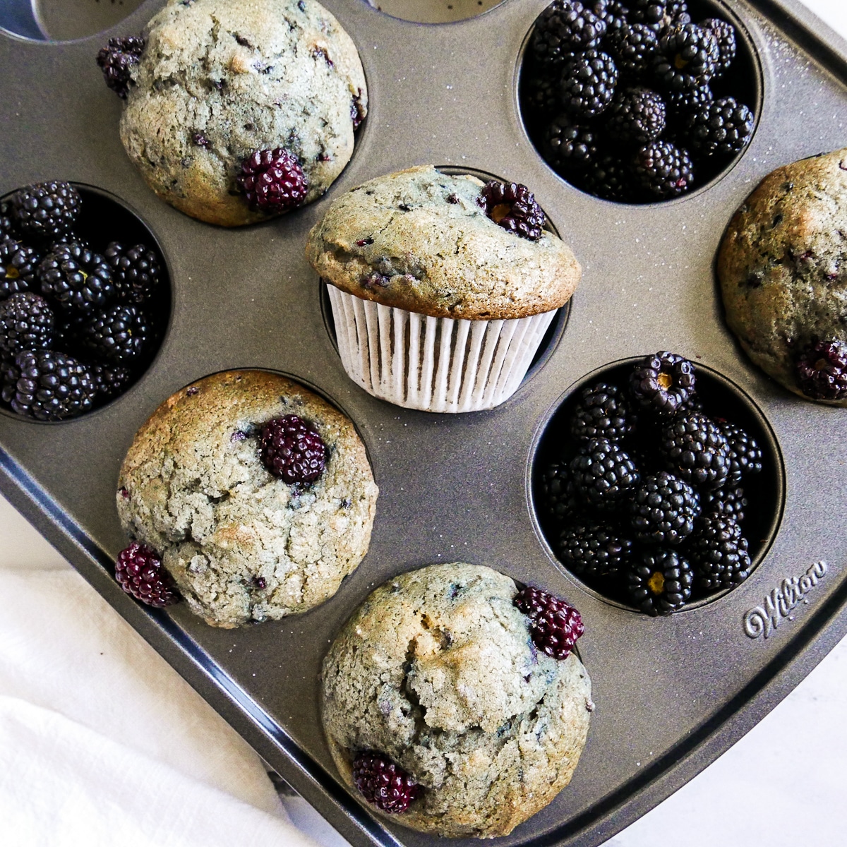 several blackberry muffins in a muffin tin with fresh blackberries.