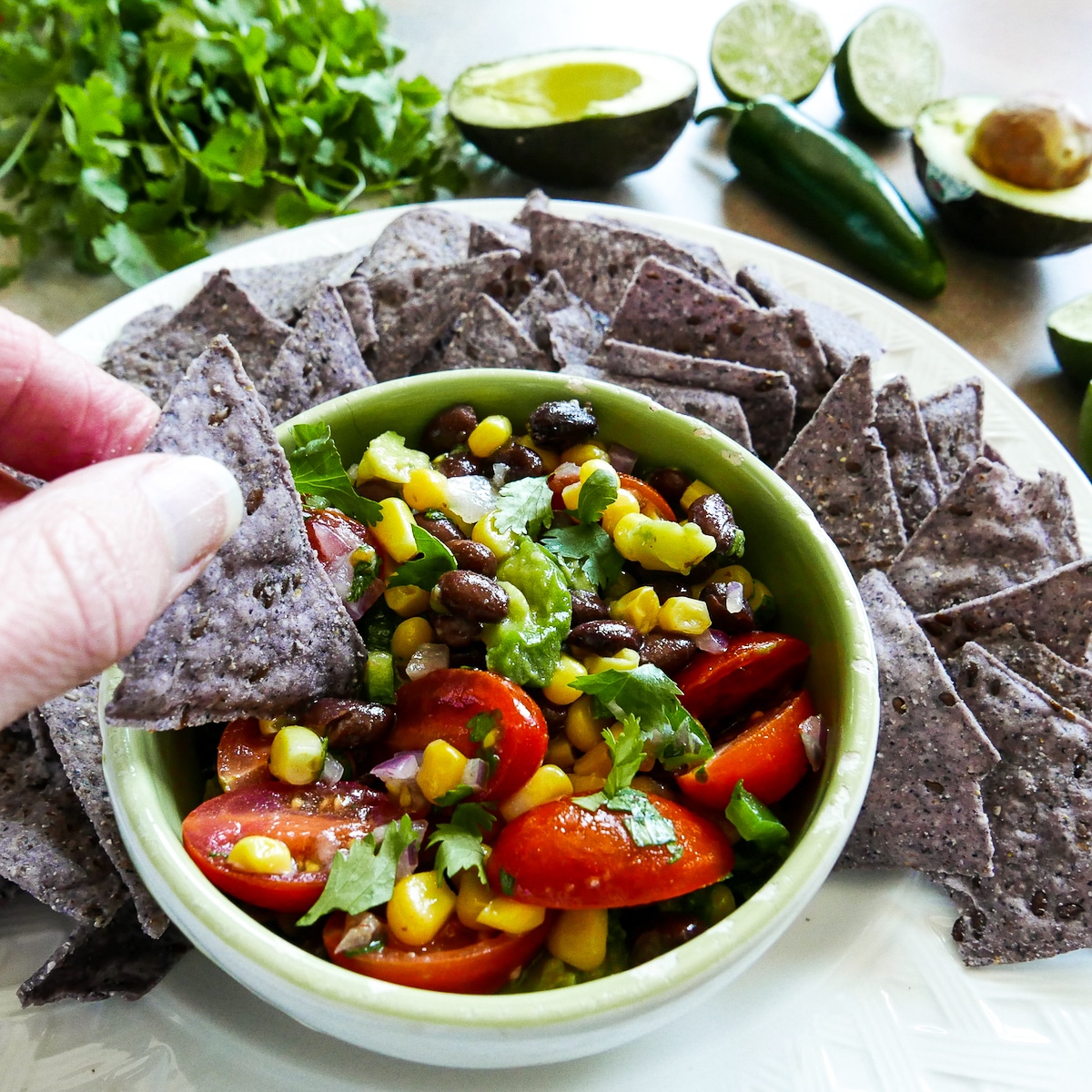 white hand dipping tortilla chip into bowl of salsa with chips on a plate.