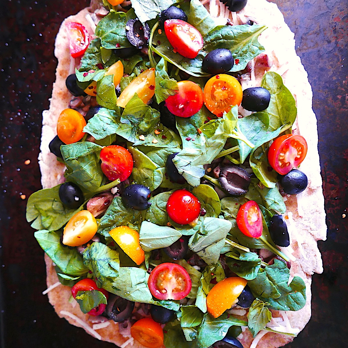 Flatbread pizza on a baking sheet topped with tomatoes, olives, and fresh basil.