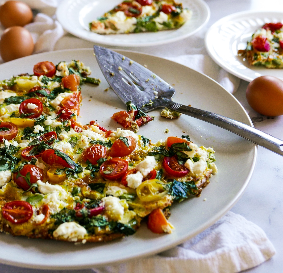 spatula-resting-on-plate-with-frittata.jpg Spatula resting on white platter holding frittata with eggs and plates in the background.