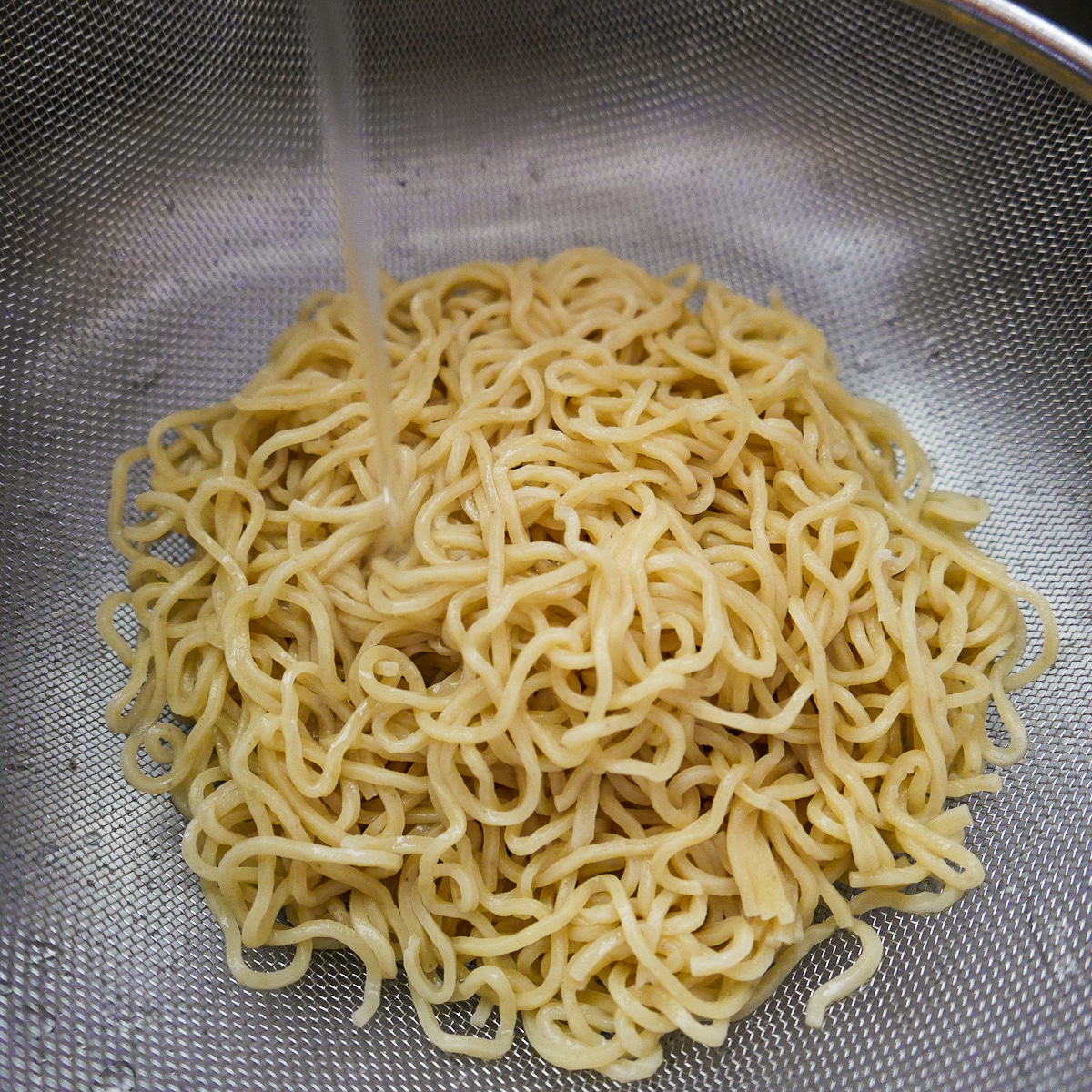 Soba noodles being rinsed under cold water in a colander. 