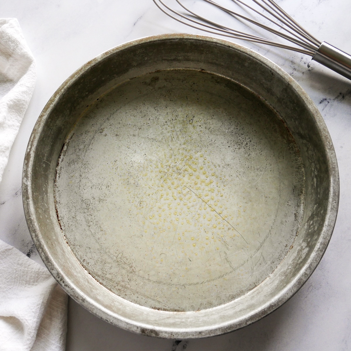 A buttered round cake pan with a whisk next to it.