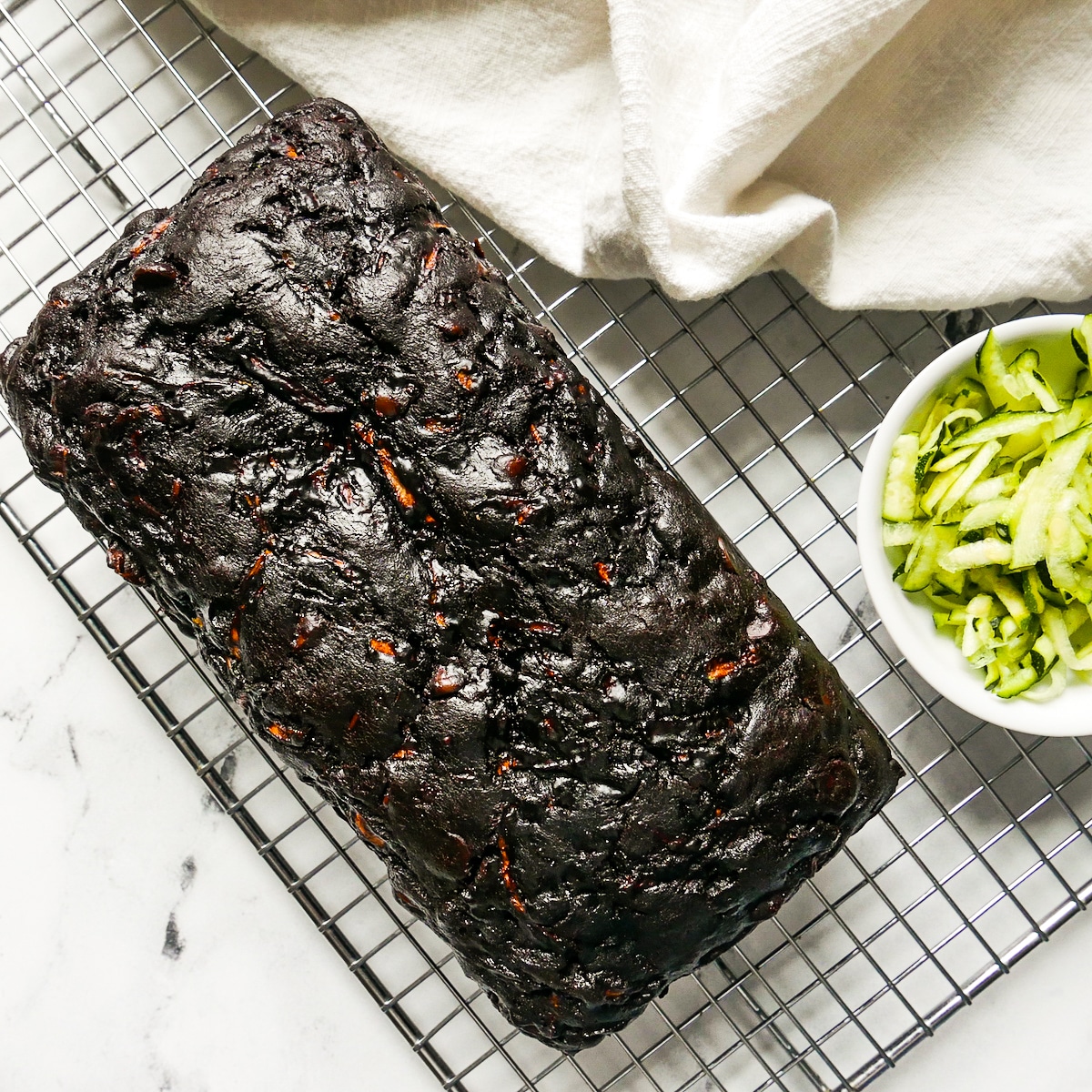 Baked bread resting on cooling rack with a cup of zucchini and white napkin.