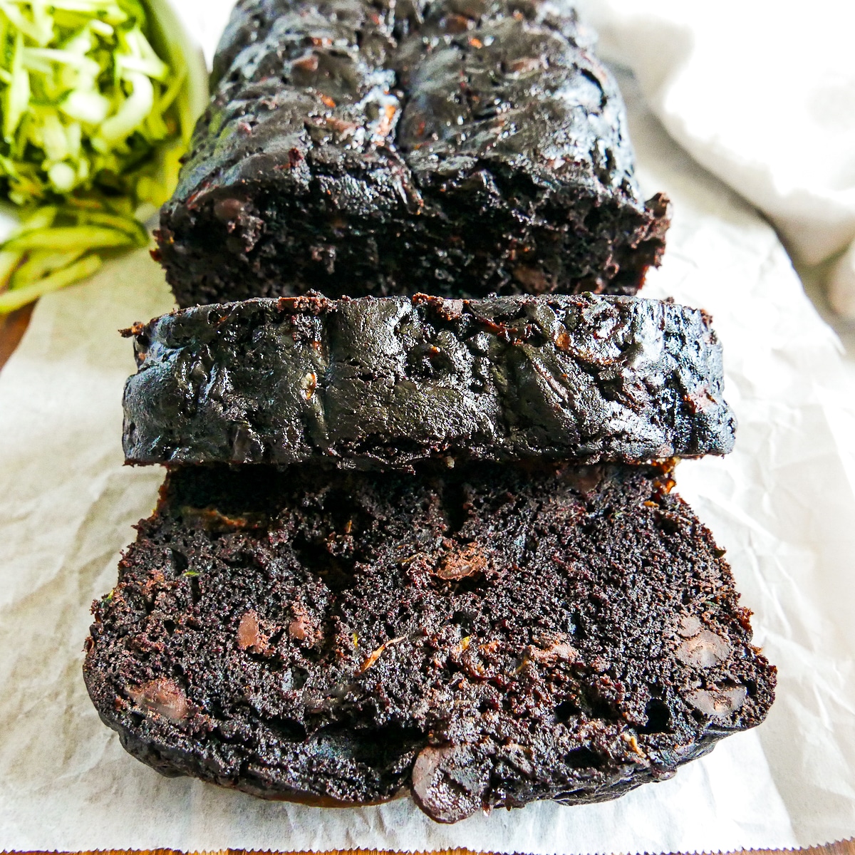 chocolate zucchini loaf sliced on parchment paper with a cup of zucchini spilling in the background.