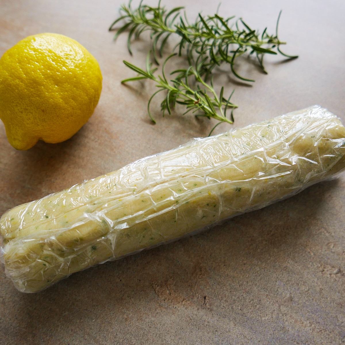 Cookie dough formed into a log and wrapped in plastic with rosemary in the background.