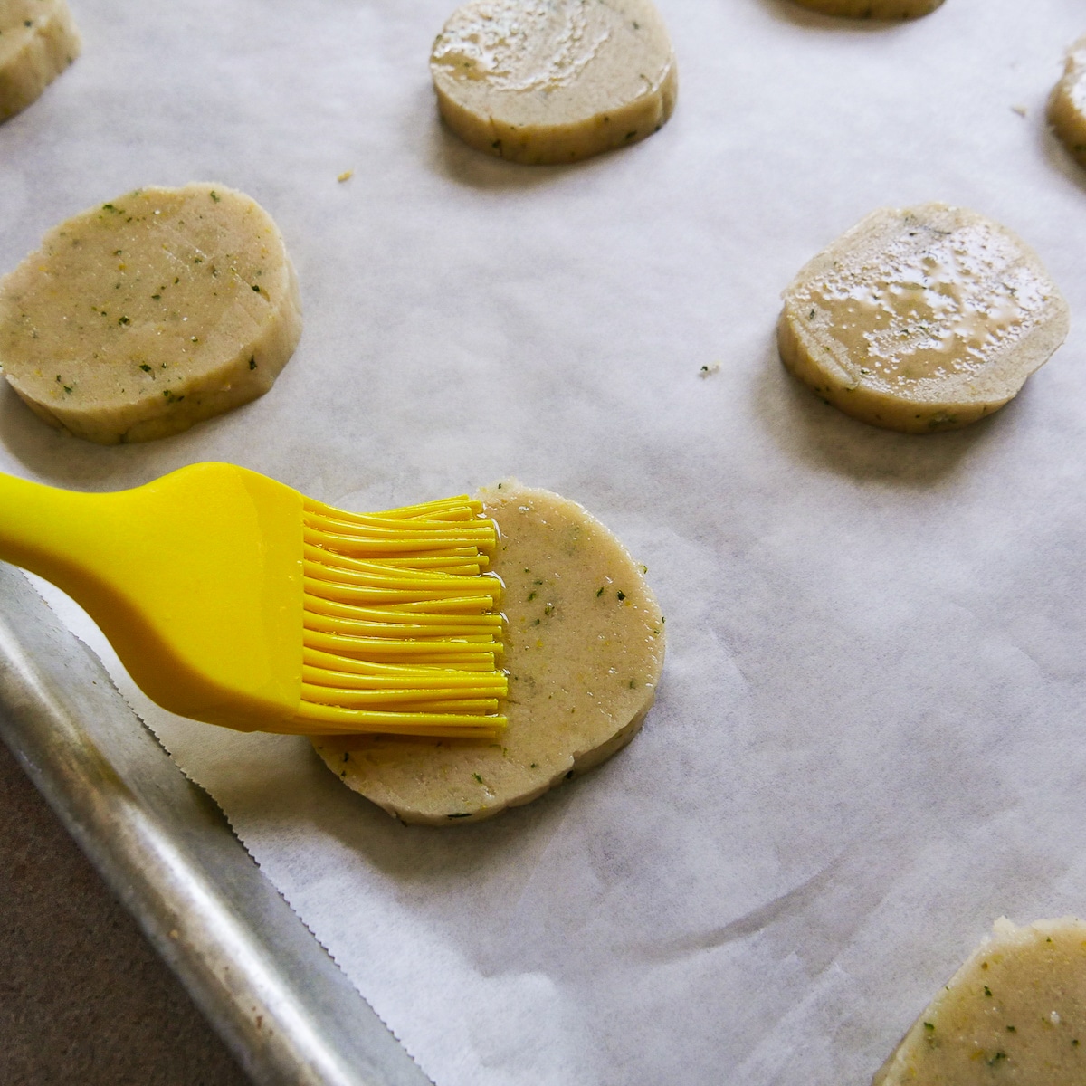 Egg white being brushed on top of sliced cookies on a baking sheet.