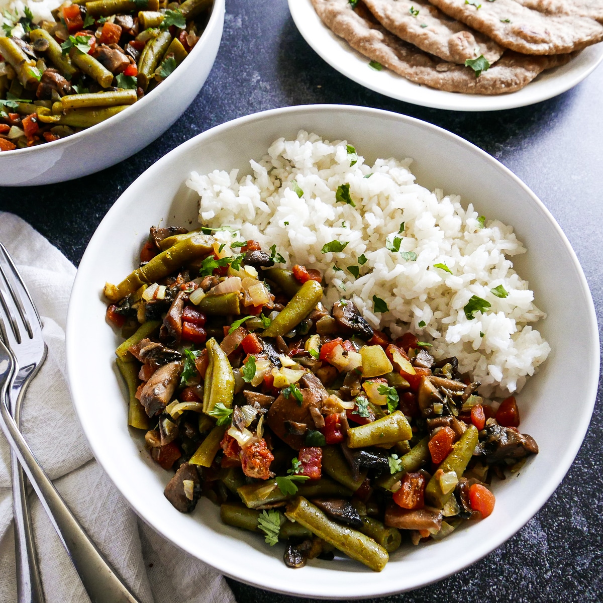 Two bowls of curried green beans with three pieces of naan on a plate.