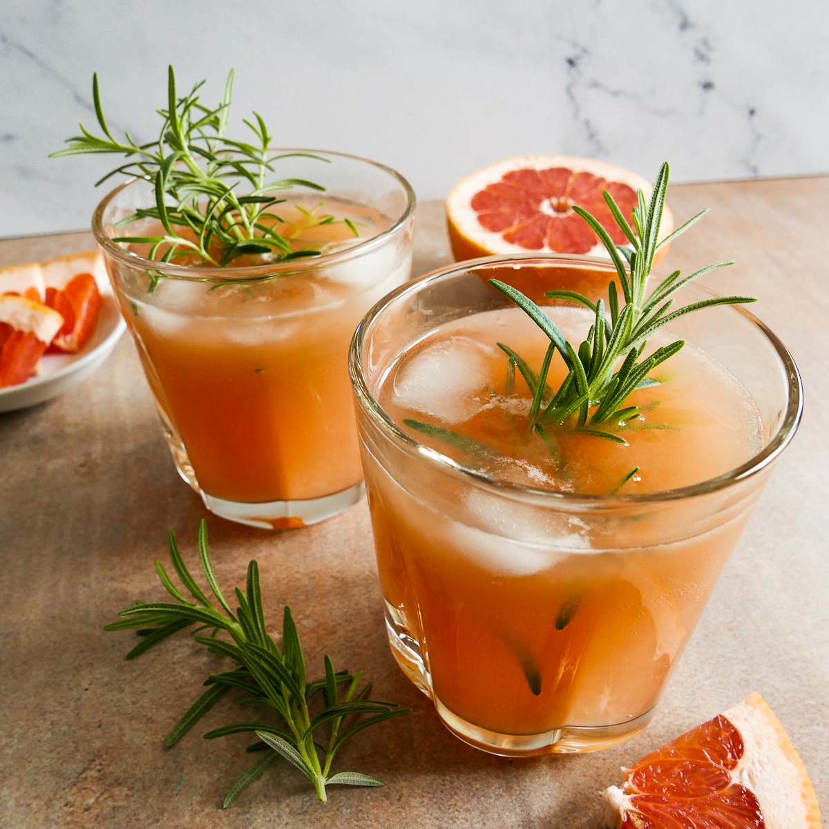 two cocktails with ice and rosemary sprigs resting on a table with sliced grapefruit nearby.