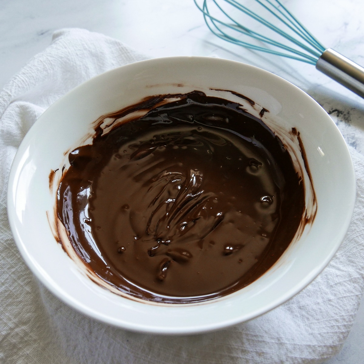 Small bowl of melted chocolate with a blue whisk and white napkin in background.