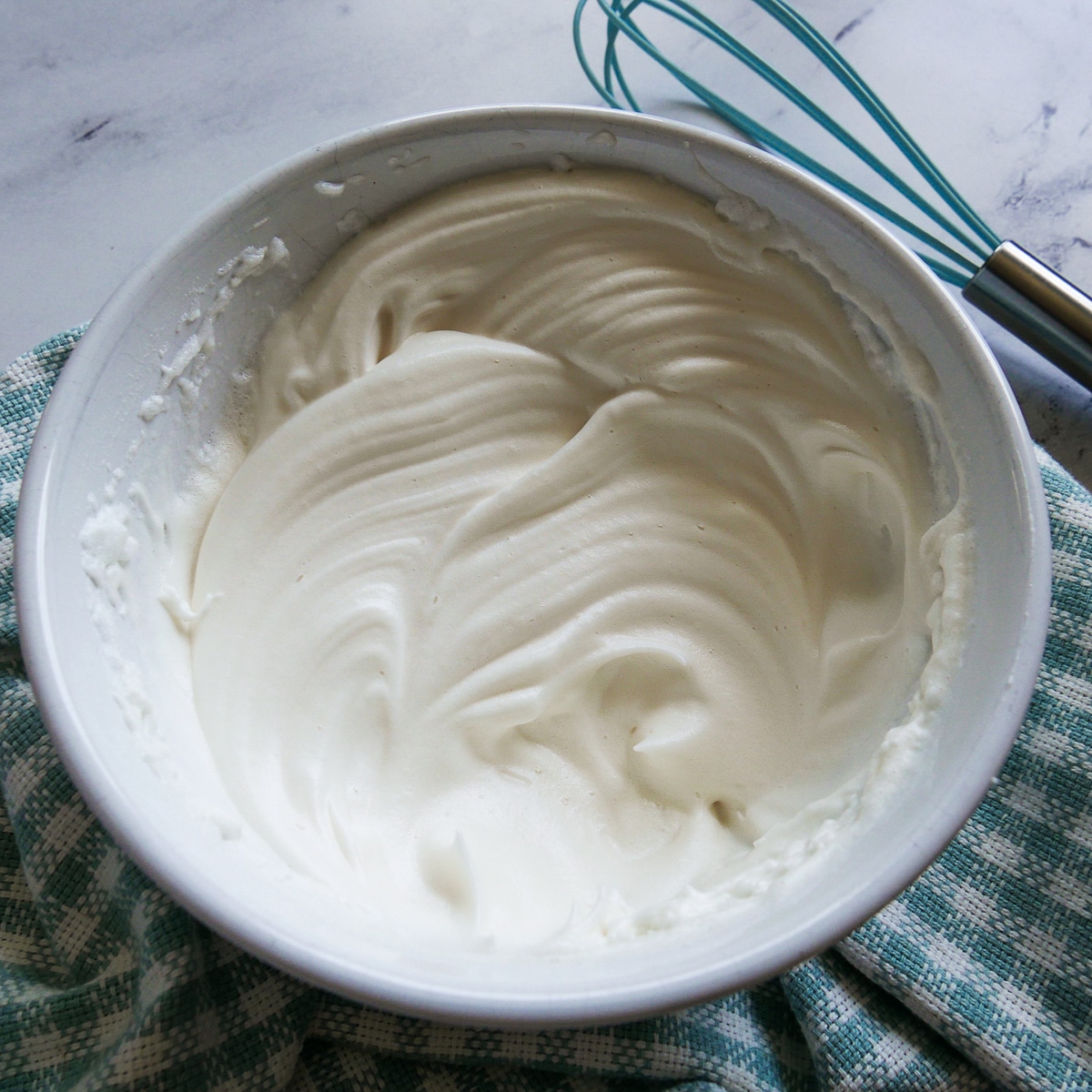 Beaten and glossy egg whites in a white bowl with a blue whisk in the background. 
