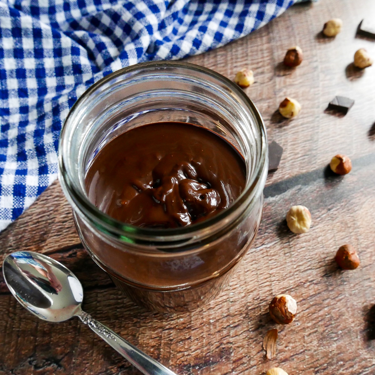 Dairy free nutella in a glass jar with a spoon and hazelnuts scattered around.