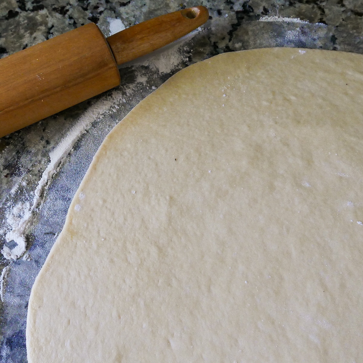 Dough rolled out onto a lightly floured surface with a rolling pin in background. 