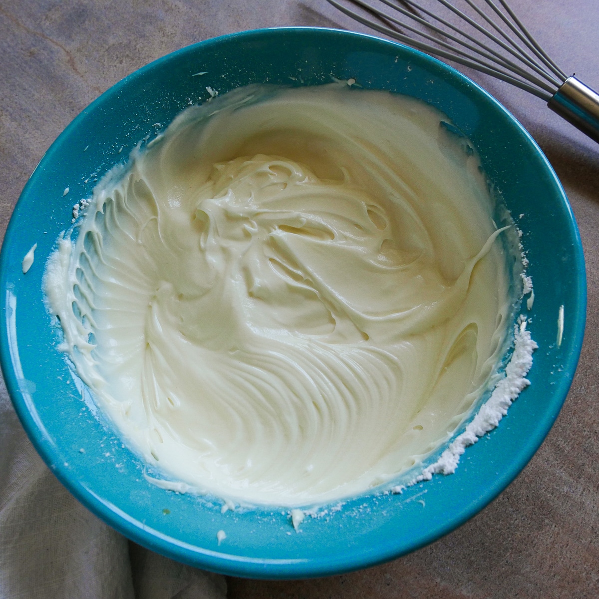 Cream cheese frosting in a blue bowl with a napkin and whisk in the background. 