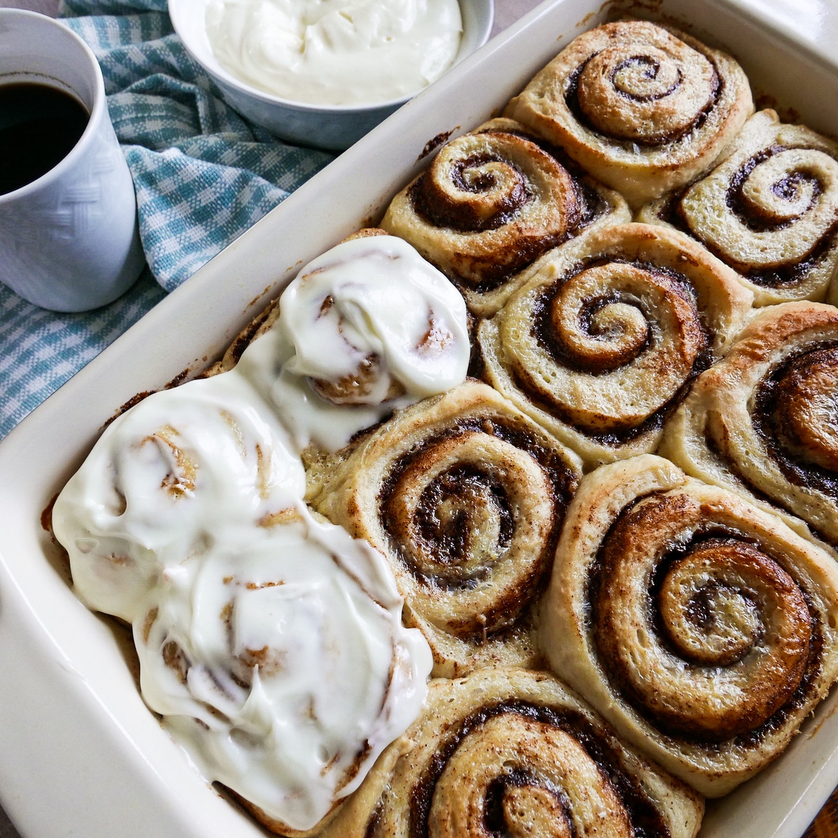 pan of partially frosted cinnamon rolls and a bowl of frosting in the background. 