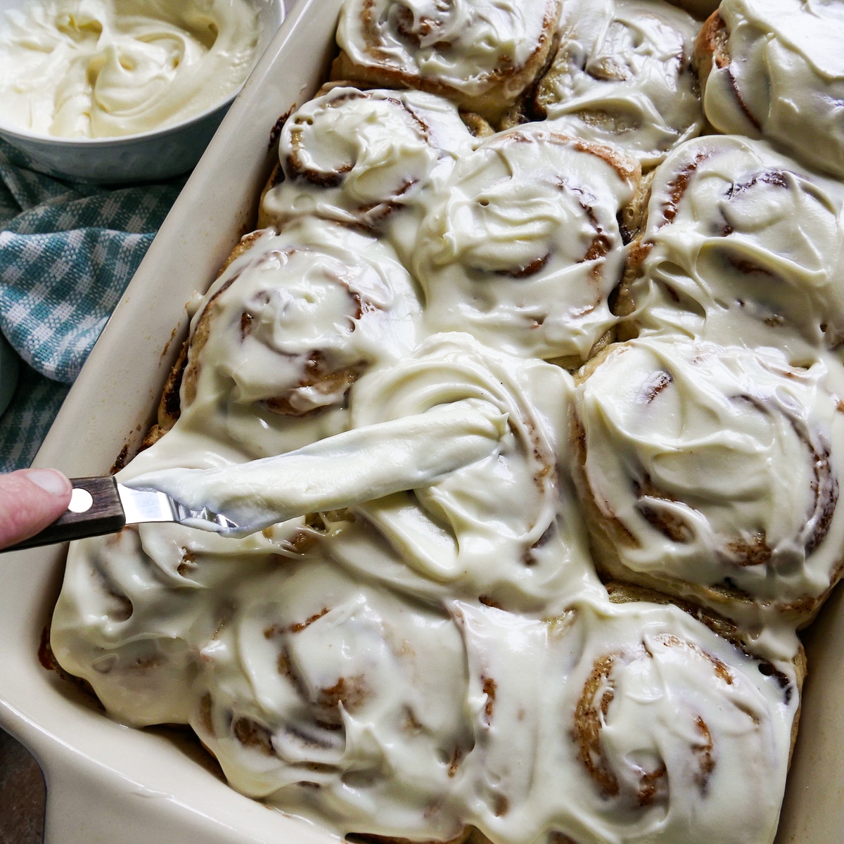 cinnamon rolls being frosted with a spatula and bowl of frosting in background. 