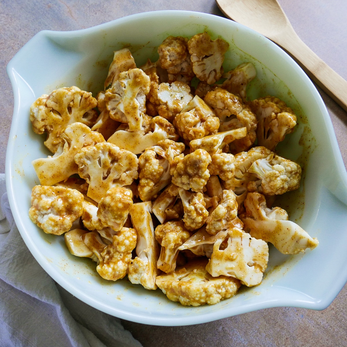 marinated cauliflower in a mixing bowl with a wooden spoon and napkin in the background. 