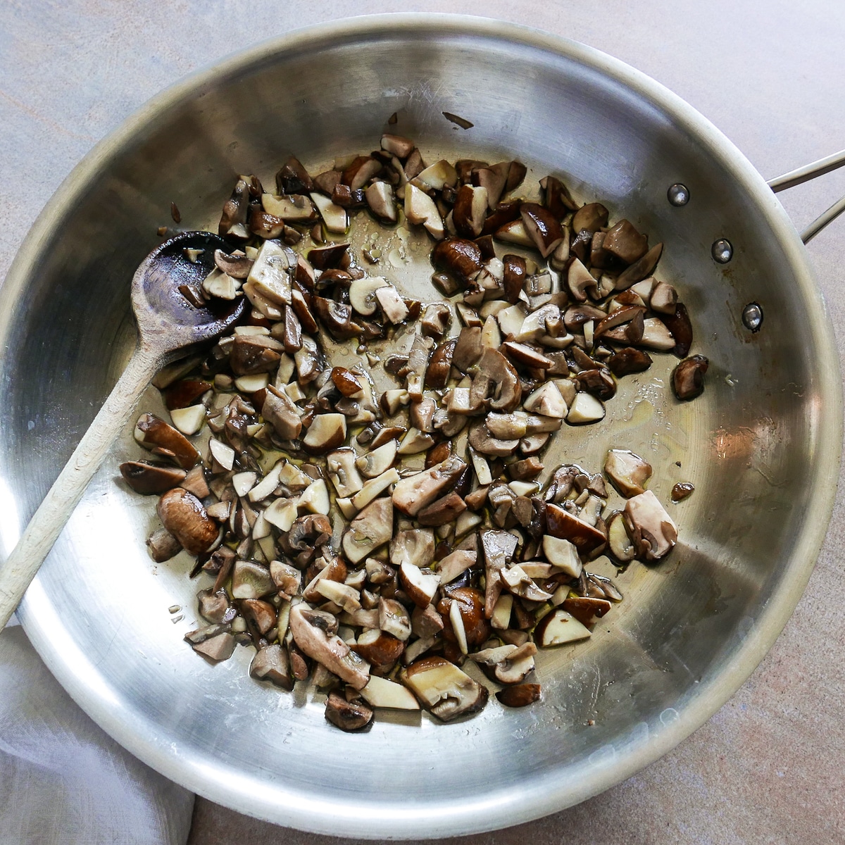 Sliced mushrooms cooking in a large skillet with a wooden spoon. 