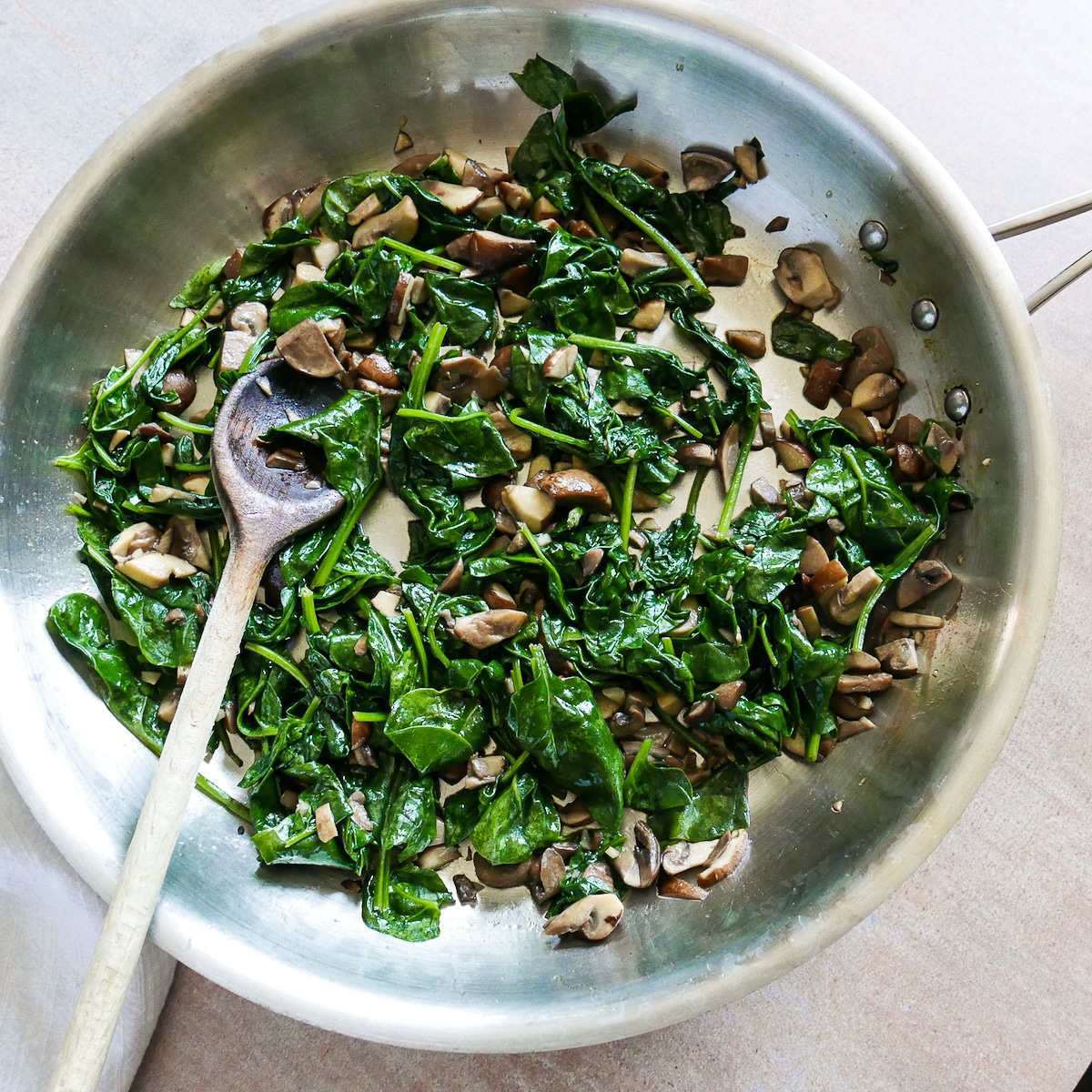 Mushrooms, garlic, and spinach cooking in a large skillet with wooden spoon.