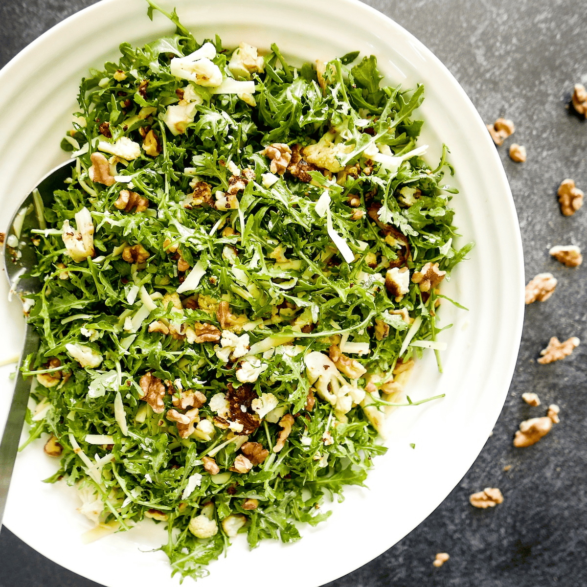 Large arugula salad with cauliflower, walnuts, and cheese with a serving spoon.