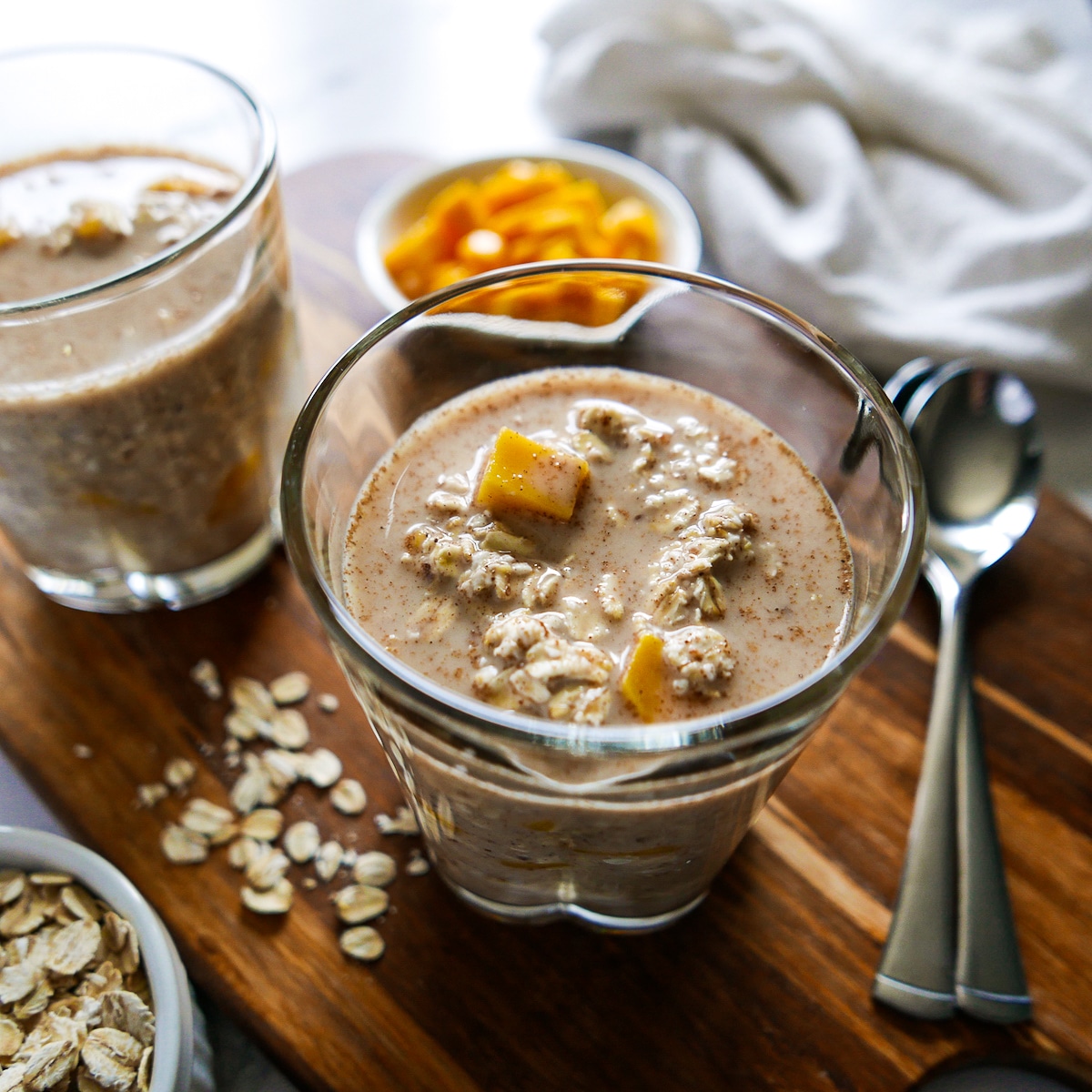 Oats mixture poured into two separate glasses with spoons and sliced mango in the background.