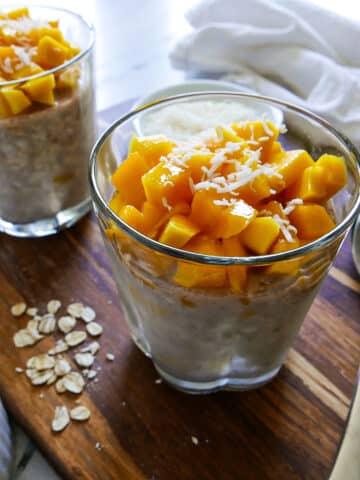 two glasses of mango overnight oats resting on a wooden cutting board.