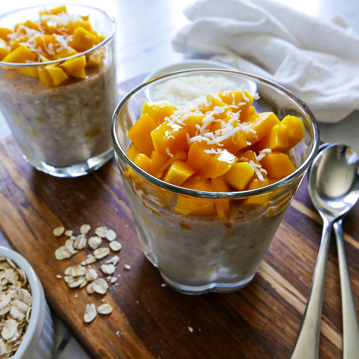 two-glasses-of-overnight-oats.jpg two glasses of mango overnight oats resting on a wooden cutting board.