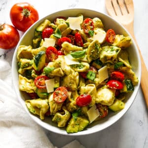 tortellini salad in a white bowl with a wooden spoon and tomatoes on the side.