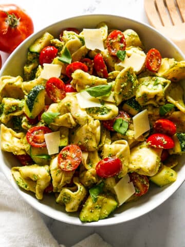 tortellini salad in a white bowl with a wooden spoon and tomatoes on the side.