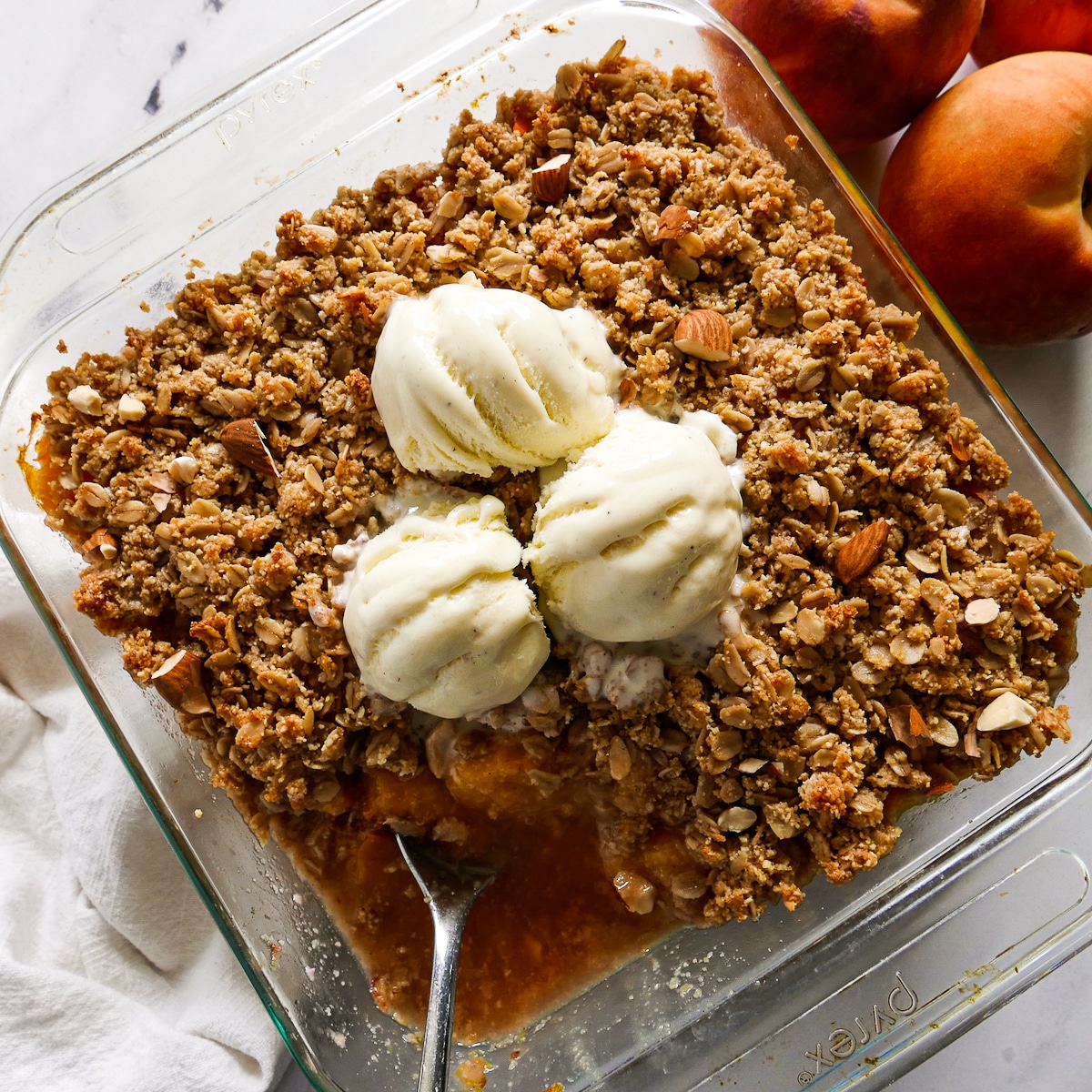 peach crisp topped with ice cream and a spoon resting in baking dish.