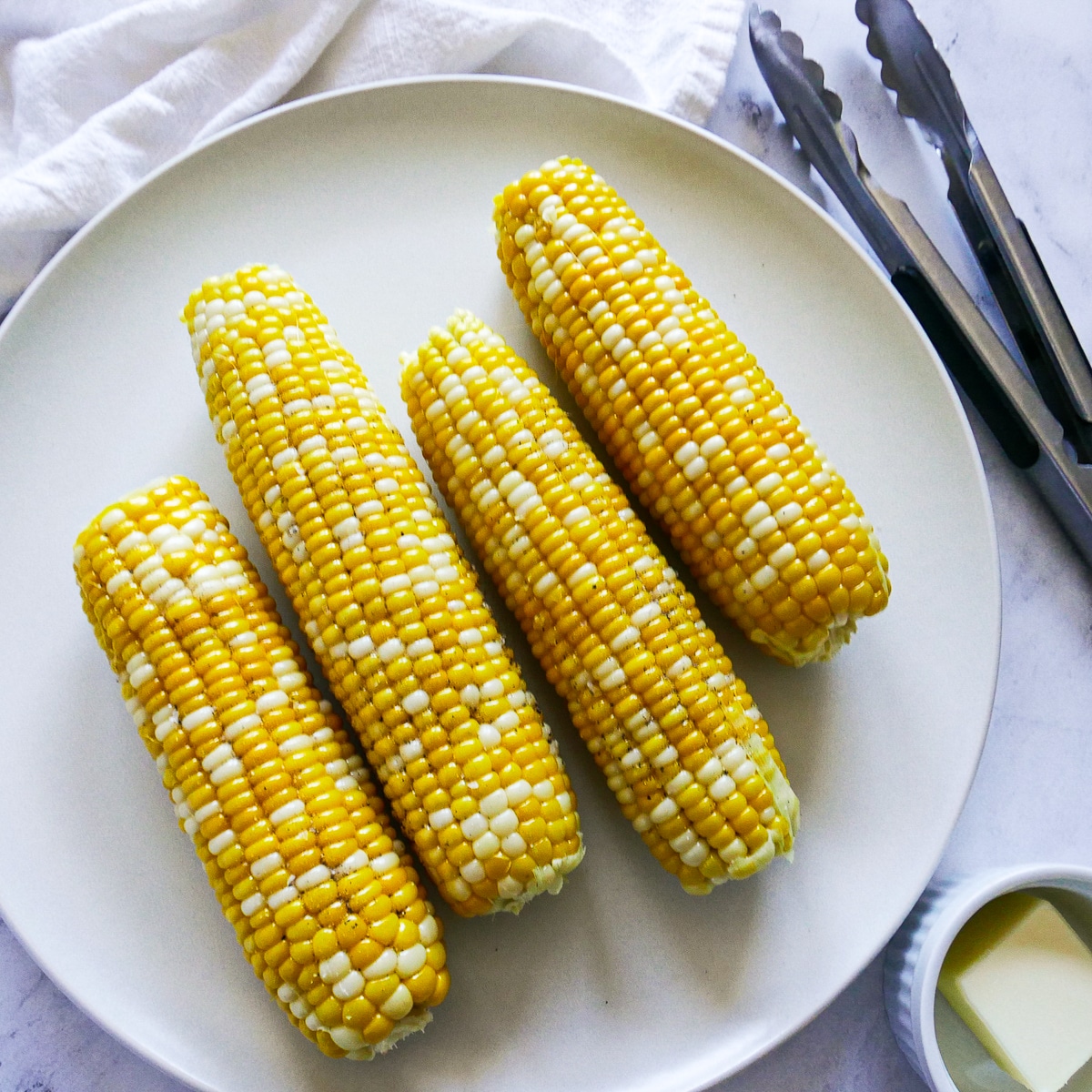 four ears of corn on a white platter with butter and tongs. 