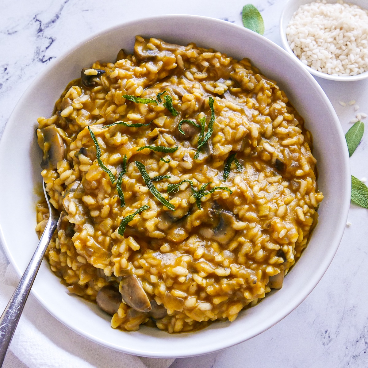 bowl of risotto with a spoon, white napkin, and sage leaves.