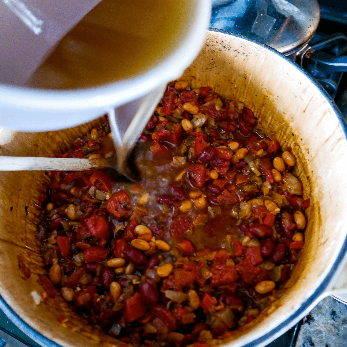 pouring vegetable broth into large pot with vegetables and beans.