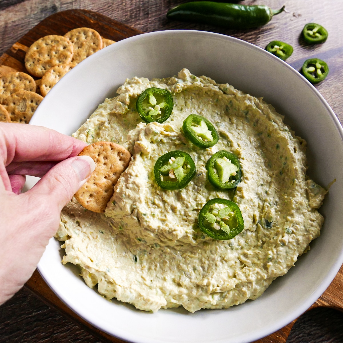 white hand dipping cracker into bowl of artichoke dip.