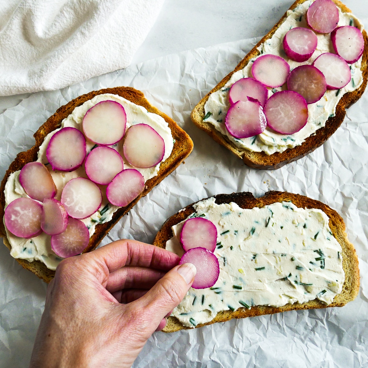 white hand arranging sliced radishes on top of toast.