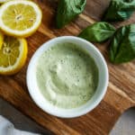 small cup of garlic basil aioli on a cutting board with lemons and basil.