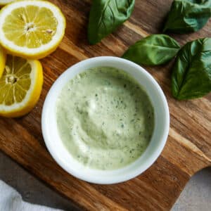 small cup of garlic basil aioli on a cutting board with lemons and basil.