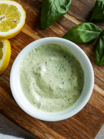 small cup of garlic basil aioli on a cutting board with lemons and basil.