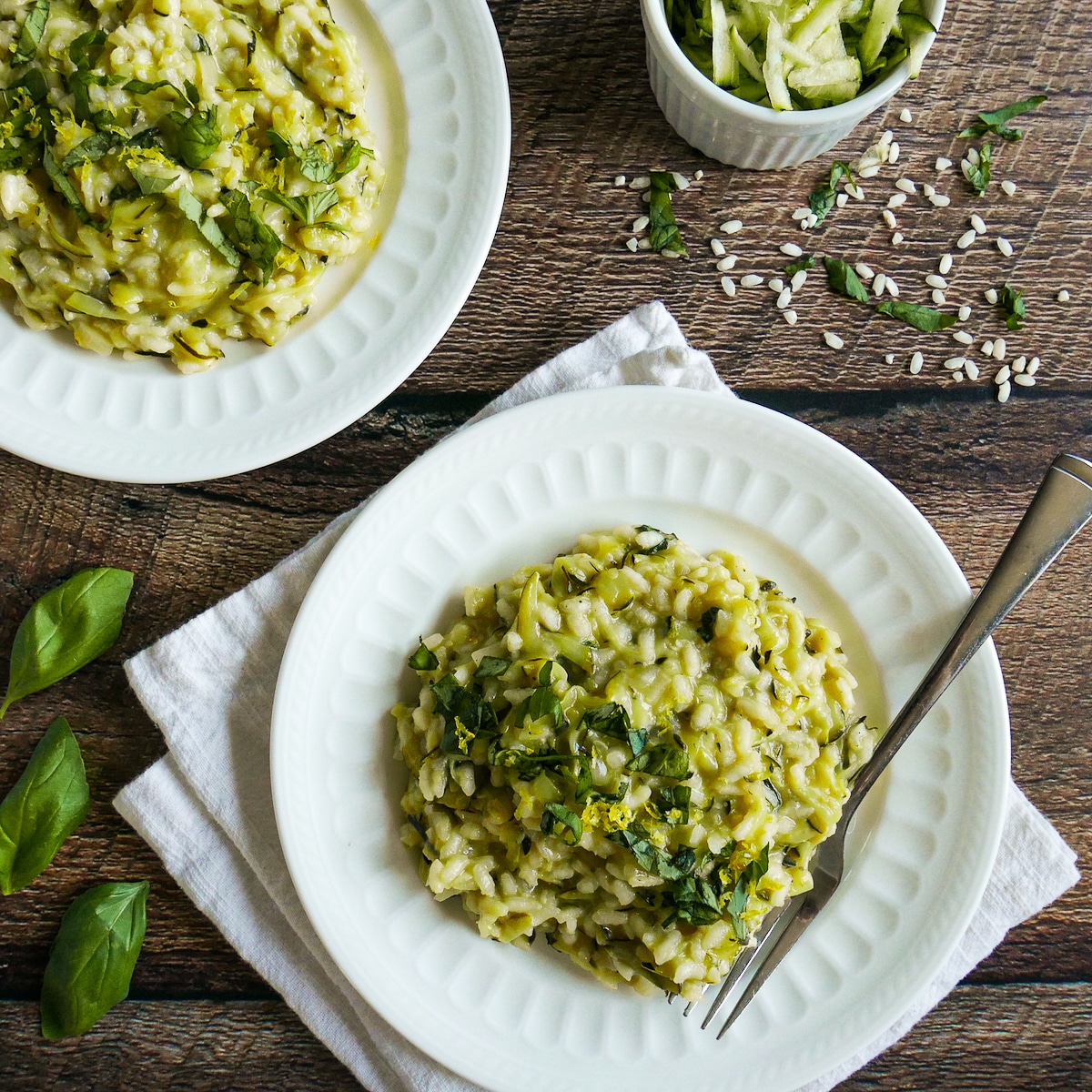 Two plates of risotto with a cup of shredded zucchini on the side.