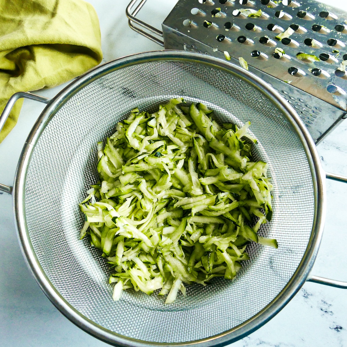 Shredded zucchini sweating in a colander resting on a bowl.