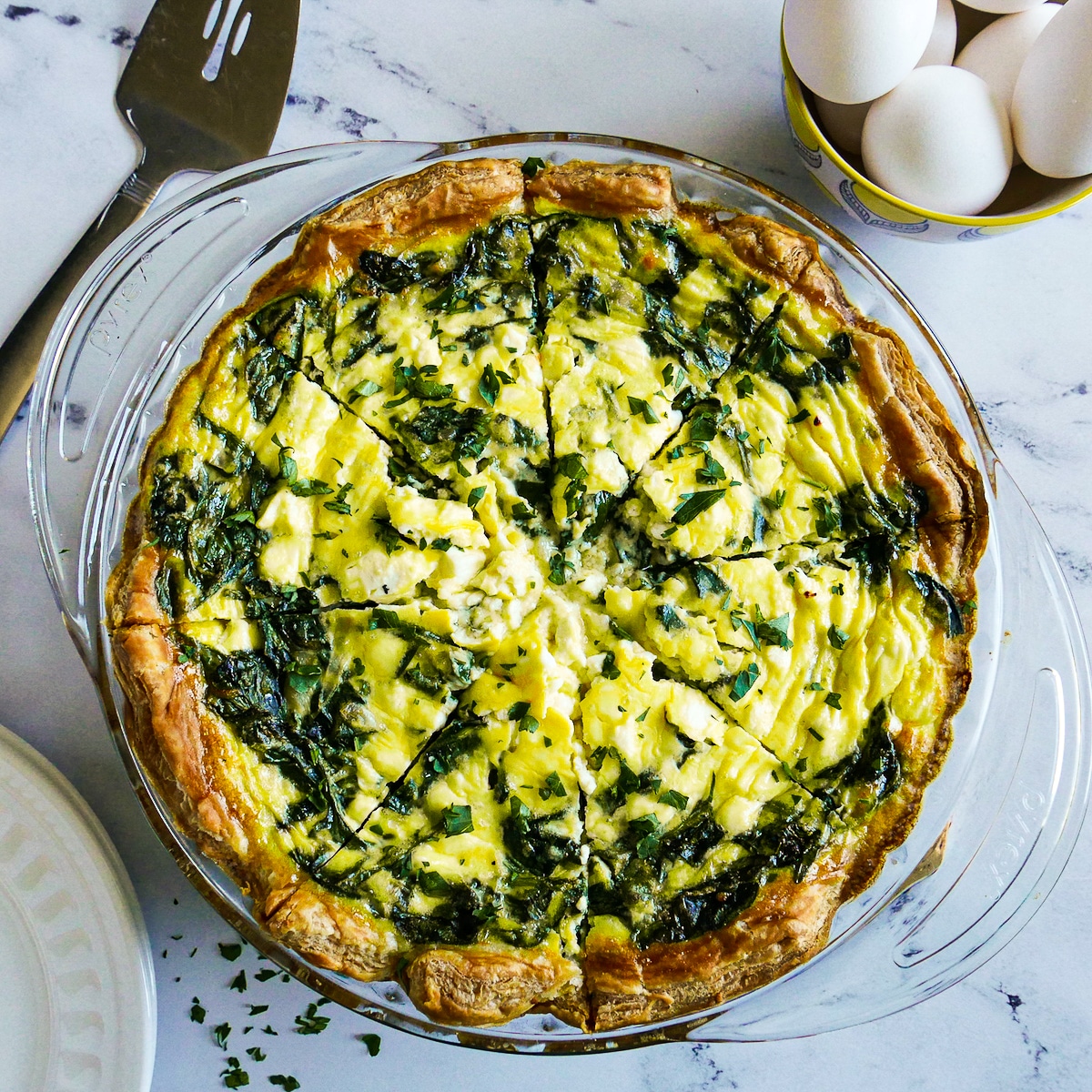 Spinach quiche cut into slices with a bowl of eggs in background.